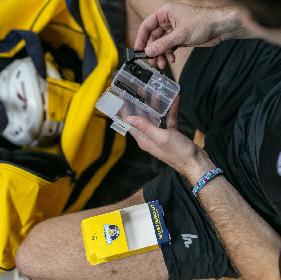 A close-up lifestyle shot of a person holding an open clear plastic case containing hockey tape and tools, with a yellow and blue package of Health Heroics hockey tape resting on their shorts. A yellow sports bag is visible in the background.